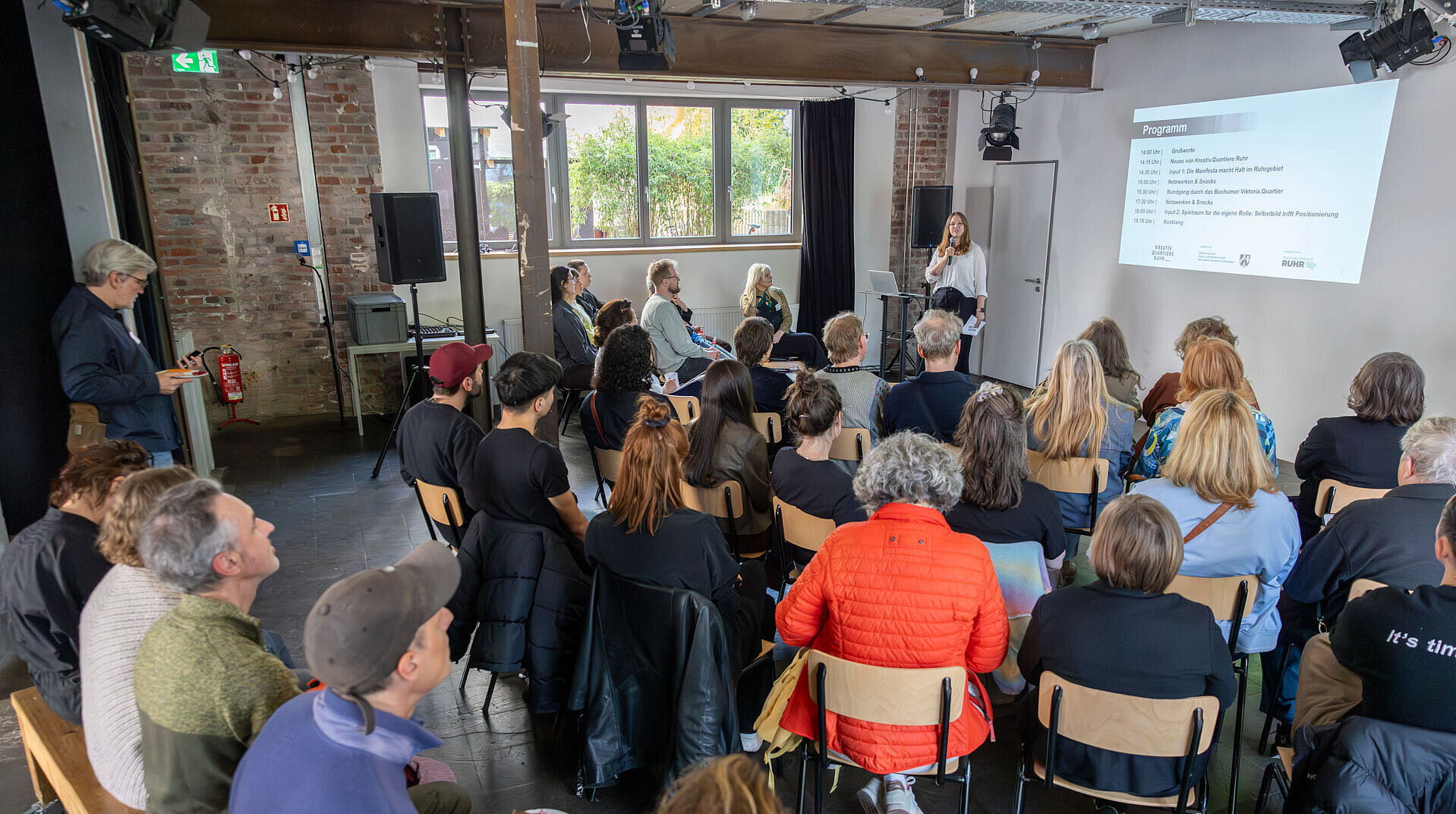 Kunst- und Kulturschaffende aus dem Netzwerk sitzen in der Quartiershalle der KoFabrik und blicken auf die Präsentation von Projektleiterin Nathalie Schraven